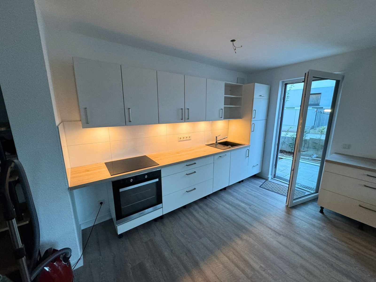 Eine kleine helle Küche mit einer Arbeitsplatte aus Holz. Auf der rechten Seite befindet sich eine geöffnete Balkontür. Die Wände der Küche sind mit hellen großen Fliesen verkleidet.