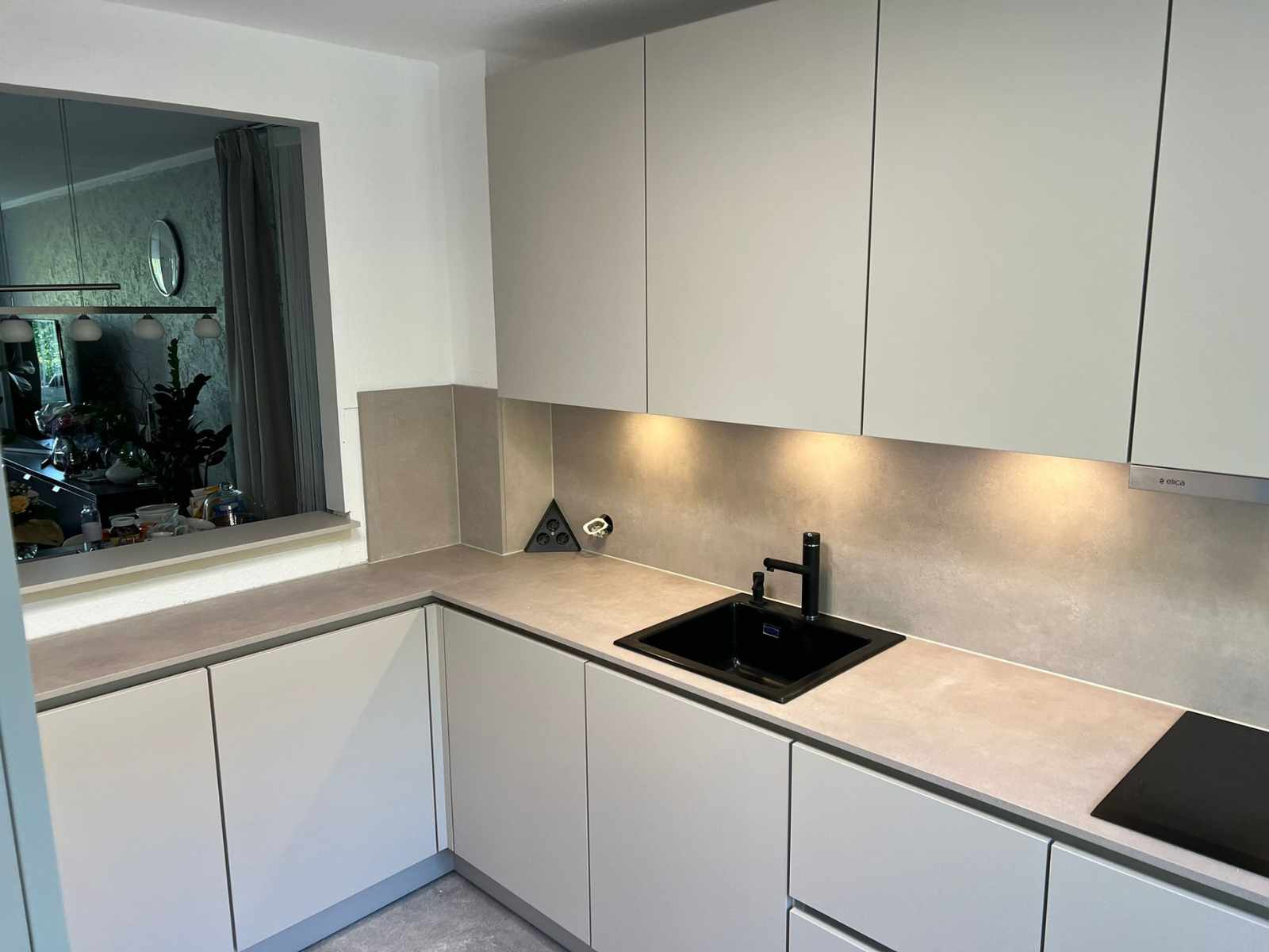 Eine helle moderne Küche mit weißen Schränken und einer hellgrauen Arbeitsplatte. Auf der linken Seite befindet sich ein großes Fenster. An der Wand an der rechten Seite sind Wandschränke montiert. Darunter moderne Beleuchtung, ein Waschbecken und ein Herd.