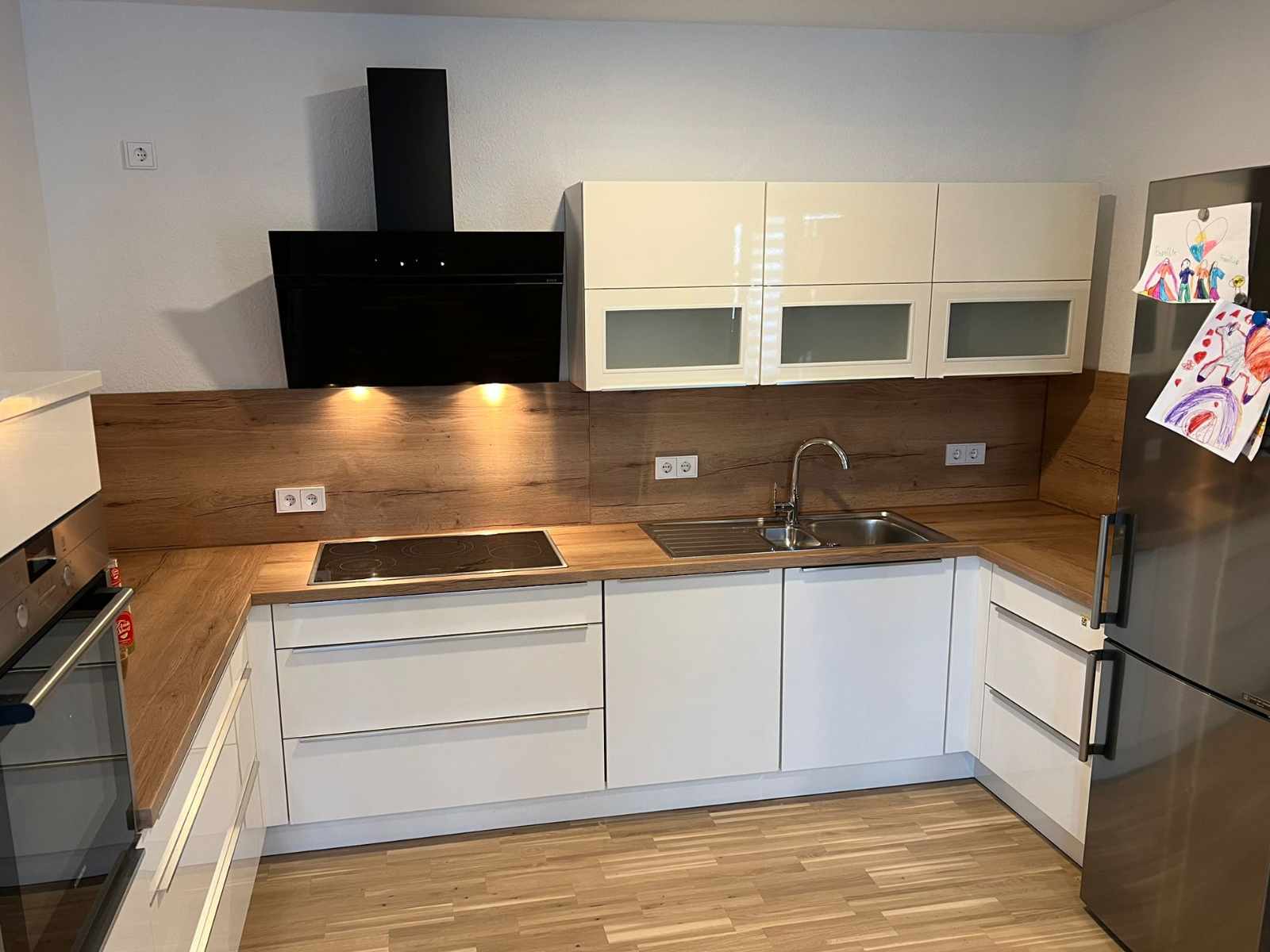 Eine neu montierte Küche, in weiß mit brauner Arbeitsplatte und brauner Wandverkleidung. Rechts ein Kühlschrank, auf welchem mal Bilder erkennt.