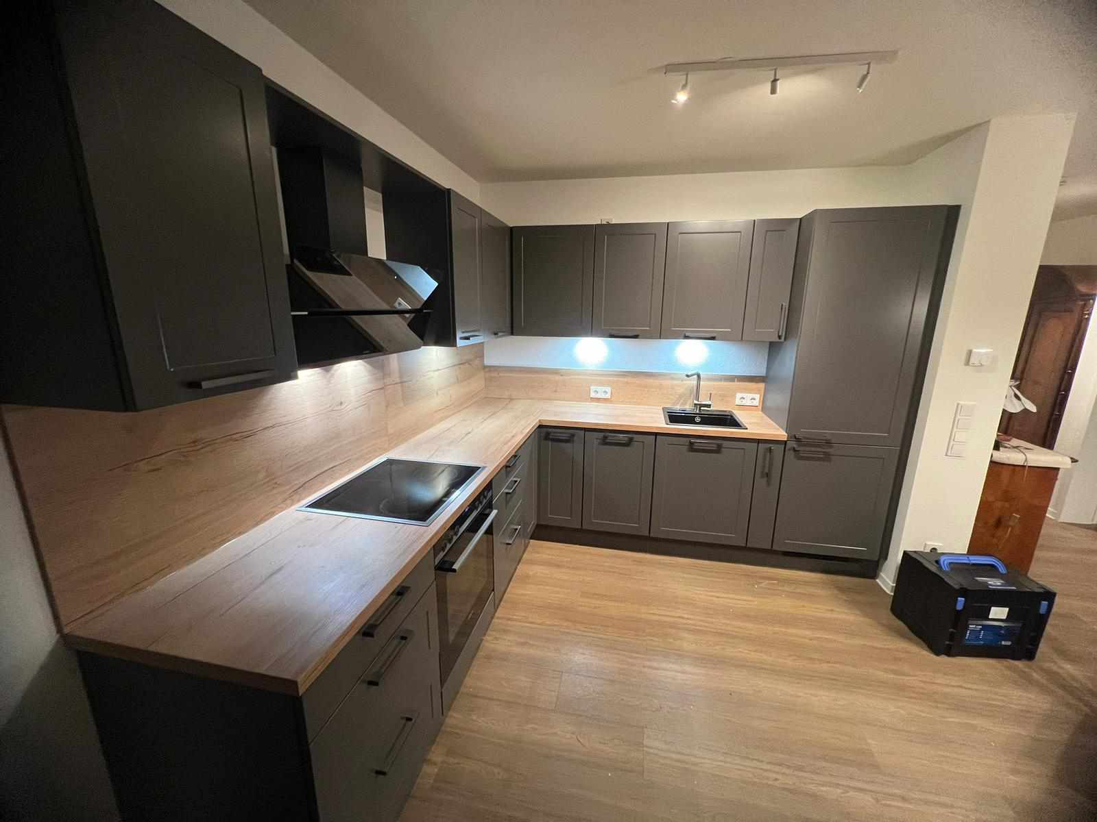 Eine grau - schwarze Küche, mit einer hellen Arbeitsplatte aus Holz. Auf de rechten Seite befindet sich ein Kühlschrank, ebenfalls grau - schwarz verkleidet. Auch die Wände sind mit dem Holz der Arbeitsplatte verkleidet.