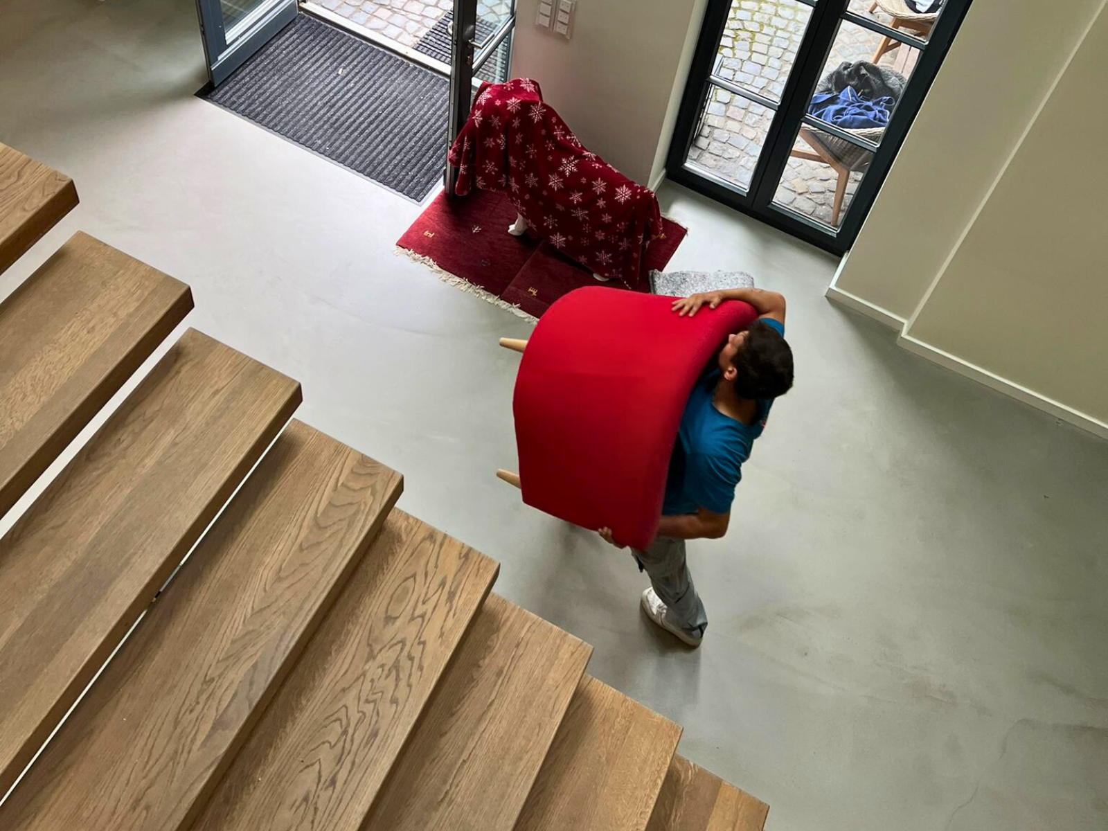 Drei Männer räumen einen gepackten Umzugstransporter aus. Sie tragen Arbeitskleidung und eine Person steht im Innenraum des Transporters, die zwei anderen befinden sich links und rechts, der geöffneten Ladefläche. Verschiedene Kartons und Möbel sind zu erkenne. Unter anderem eine Staffelei.