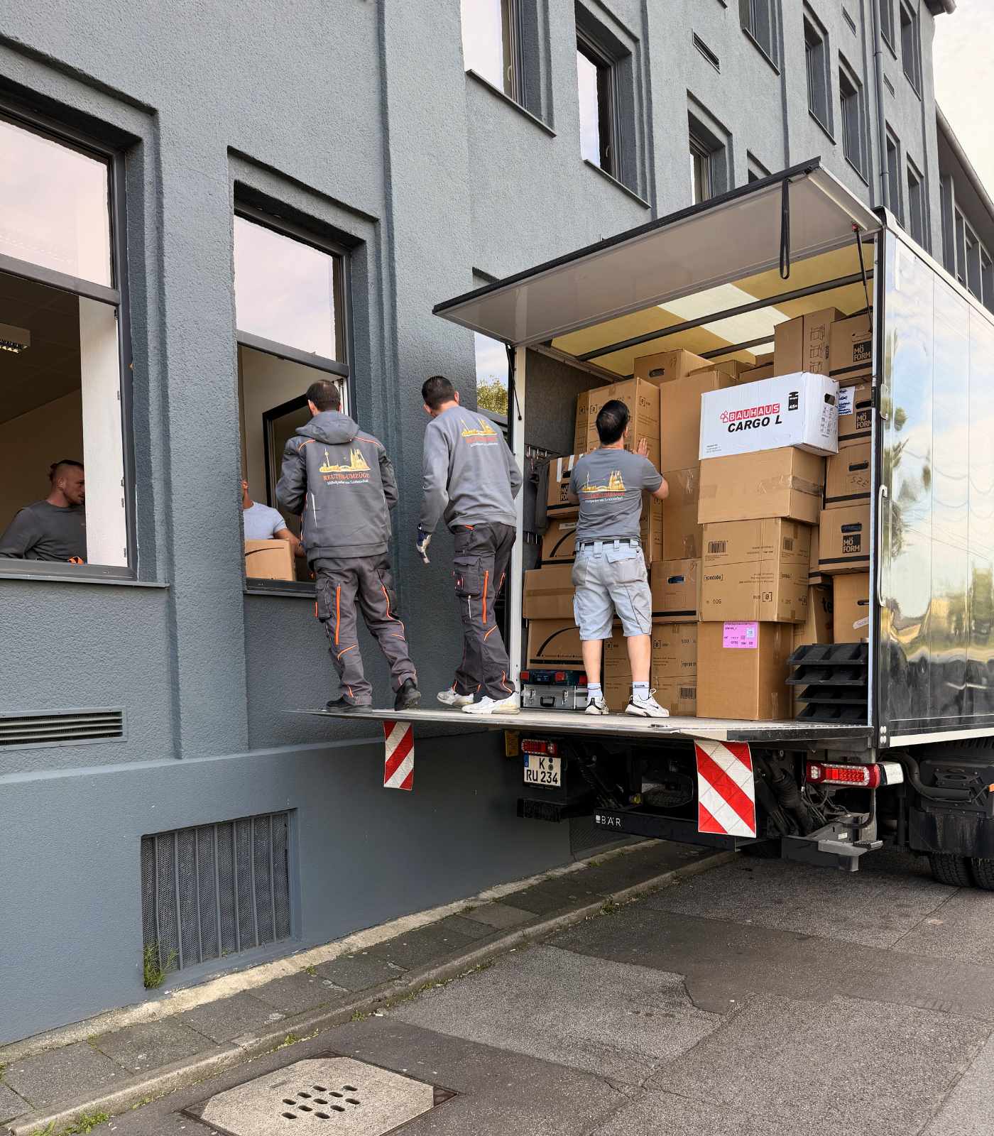 Mehrere Mitarbeiter von Reuter Umzüge beladen einen Umzugs-Lkw mit Kartons vor einem Bürogebäude in Köln.