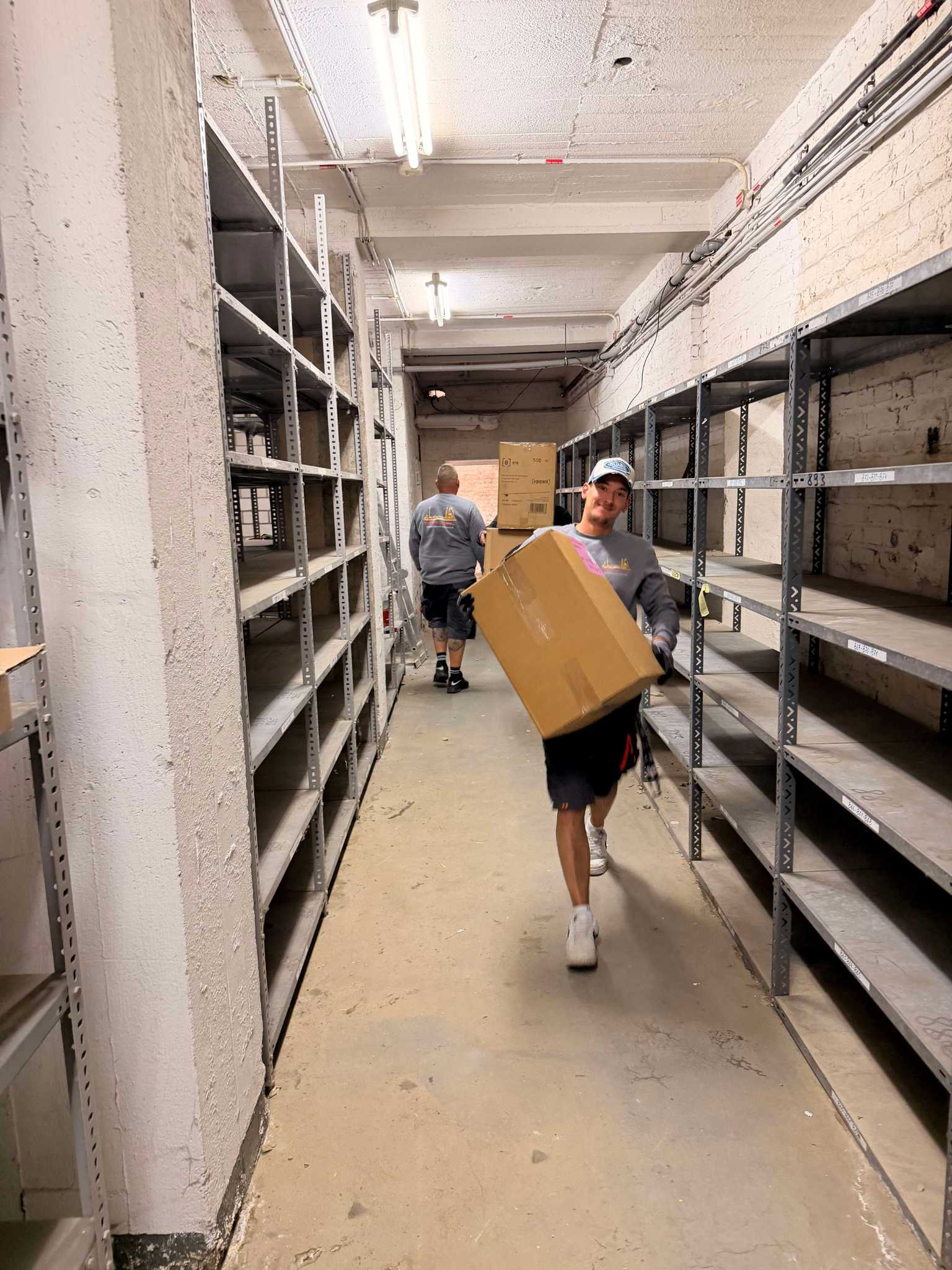 Mitarbeiter von Reuter Umzüge räumen im Rahmen eines Büroumzugs in Köln Lagerflächen aus und transportieren Umzugskartons durch einen Regalgang.